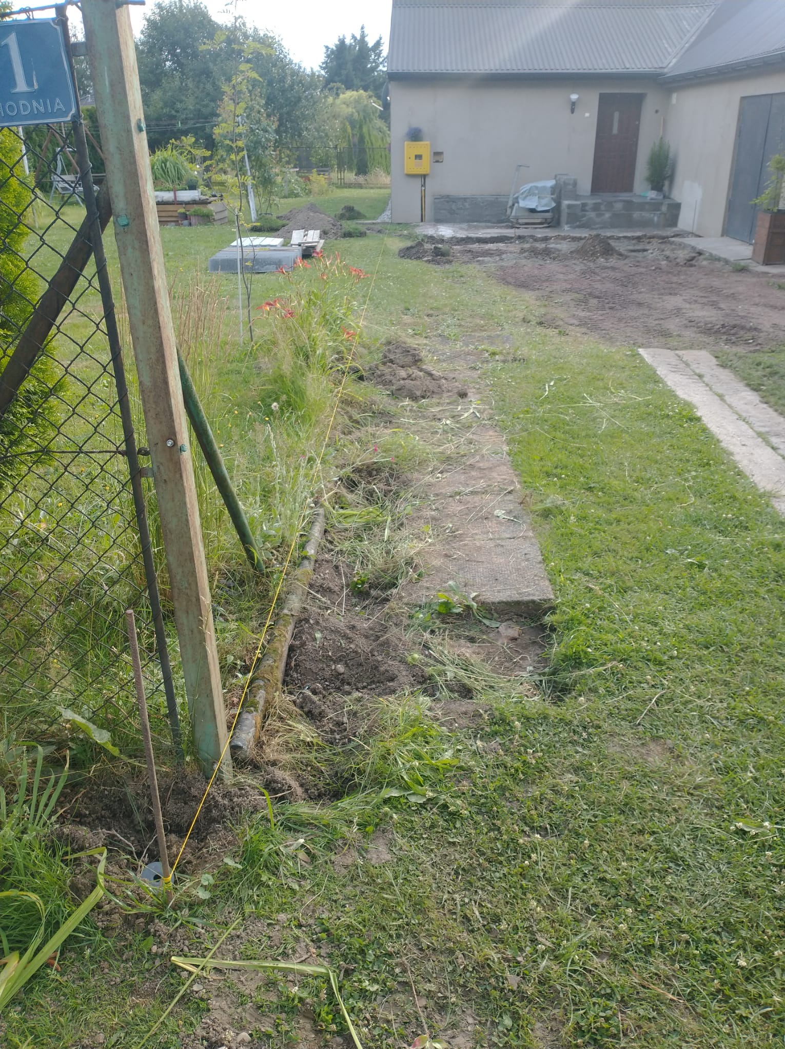 Przygotowanie terenu pod ścieżkę w ogrodzie: wytyczona linia, częściowo usunięta darń, widoczny fragment ogrodzenia i domu w tle. Prace ziemne w toku.