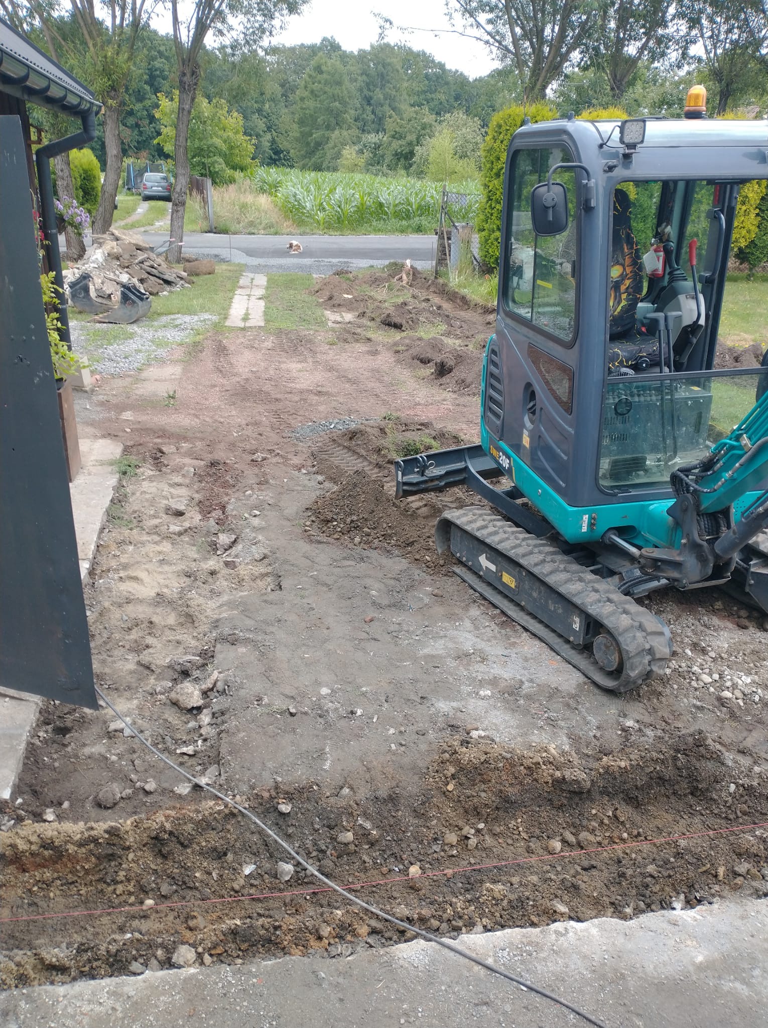 Mini koparka na posesji w trakcie prac ziemnych, widoczne ślady gąsienic i wykop. W tle droga i zieleń. Prace przygotowawcze do dalszych etapów budowy.