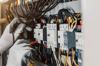 Elektryk w rękawicach ochronnych mierzy napięcie w rozdzielnicy elektrycznej za pomocą multimetru. Widoczne przewody i zabezpieczenia.