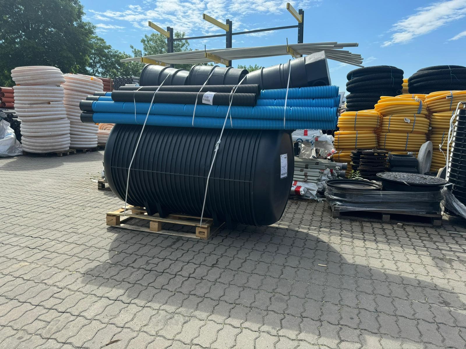 Czarny zbiornik na palecie, otoczony rurami drenażowymi w kolorach: białym, niebieskim, czarnym i żółtym. Widoczne elementy studzienek i inne akcesoria.