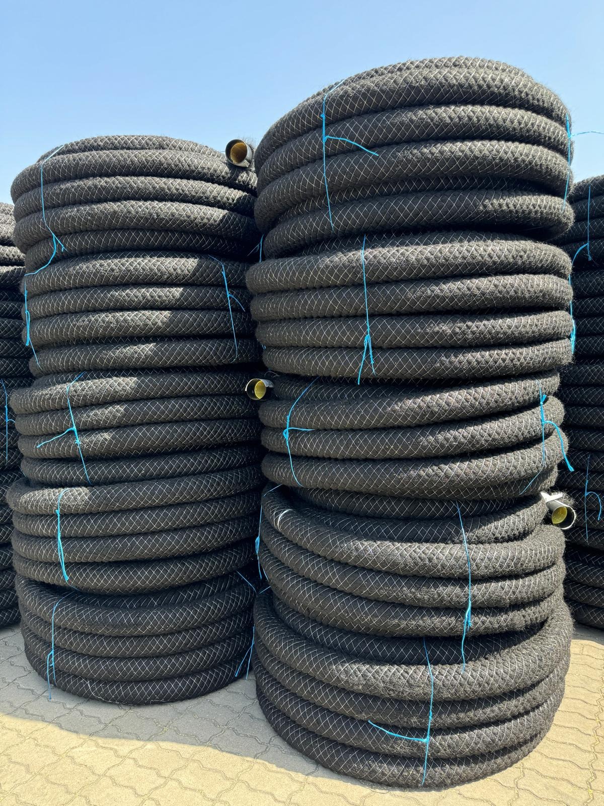 Stosy czarnych rur drenarskich z niebieskim sznurkiem na zewnątrz, ułożone na betonowej powierzchni pod błękitnym niebem. Widoczne końcówki rur.