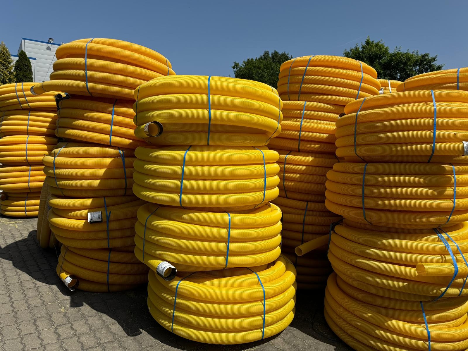 Stosy żółtych, karbowanych rur drenarskich, spiętych niebieską taśmą, ułożone na szarym bruku. Widoczne etykiety na niektórych rolkach. Słoneczny dzień.