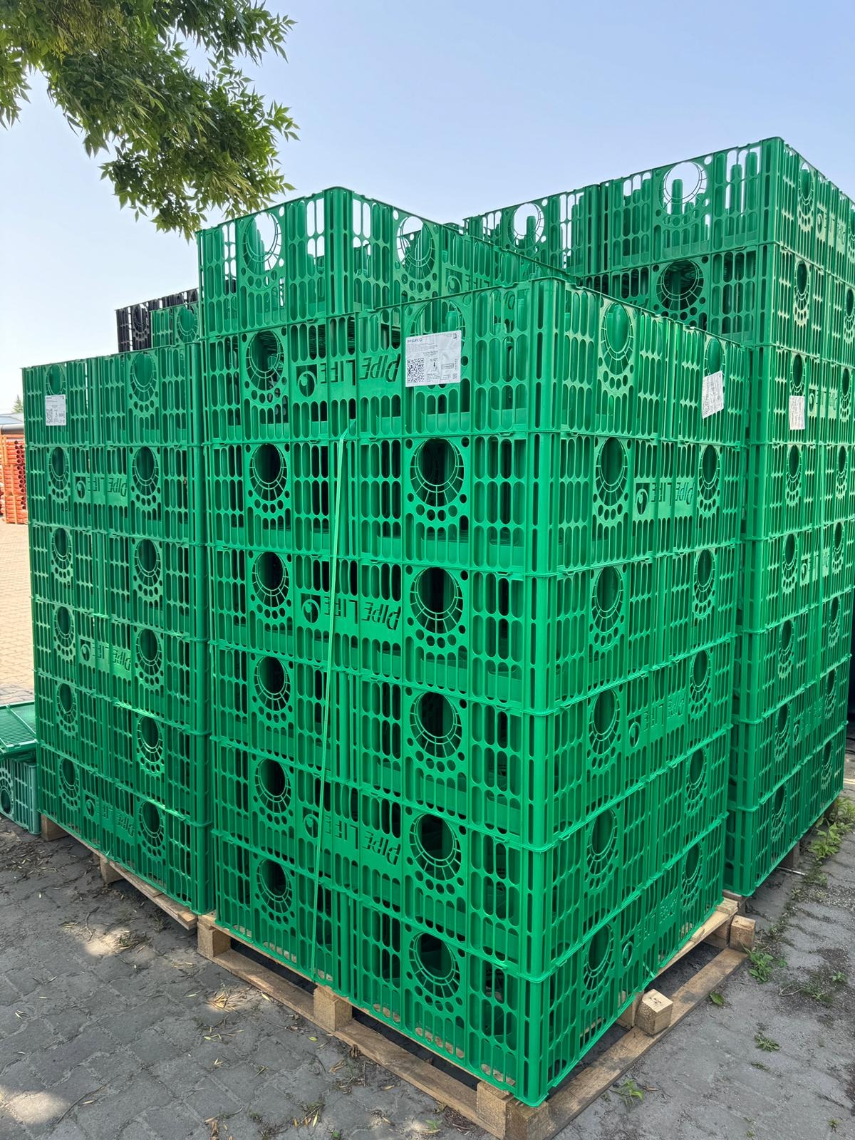 Stos zielonych, ażurowych skrzynek z tworzywa sztucznego na palecie, z widocznym napisem 'PIPE LIFE', na tle kostki brukowej i fragmentu drzewa.