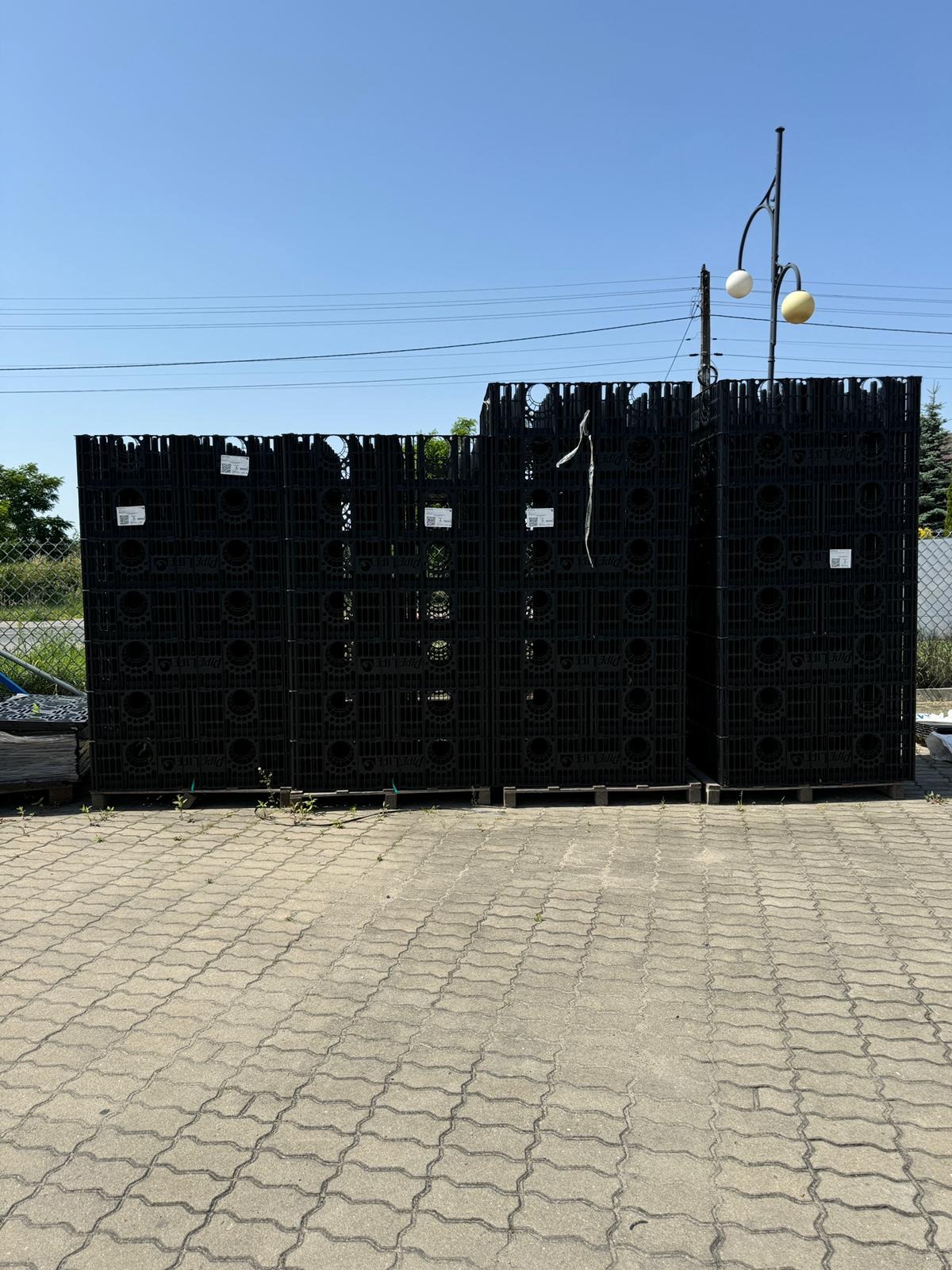 Czarne, ażurowe moduły składowane na paletach, prawdopodobnie elementy systemów odwodnień lub oczyszczalni, na tle błękitnego nieba i zieleni. Widoczne słupy oświetleniowe.