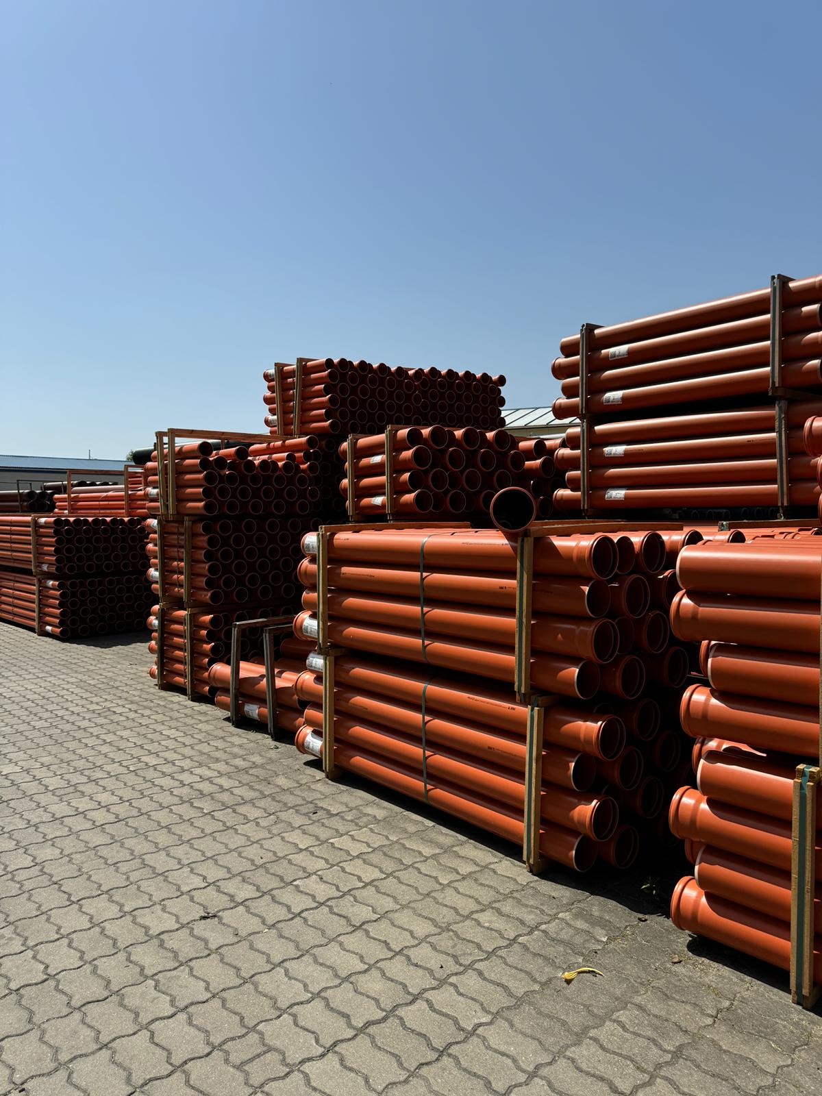 Pomarańczowe rury kanalizacyjne ułożone w stosy na paletach, widoczne na zewnątrz w słoneczny dzień. Różne średnice rur, regularny układ, szare podłoże z kostki brukowej.