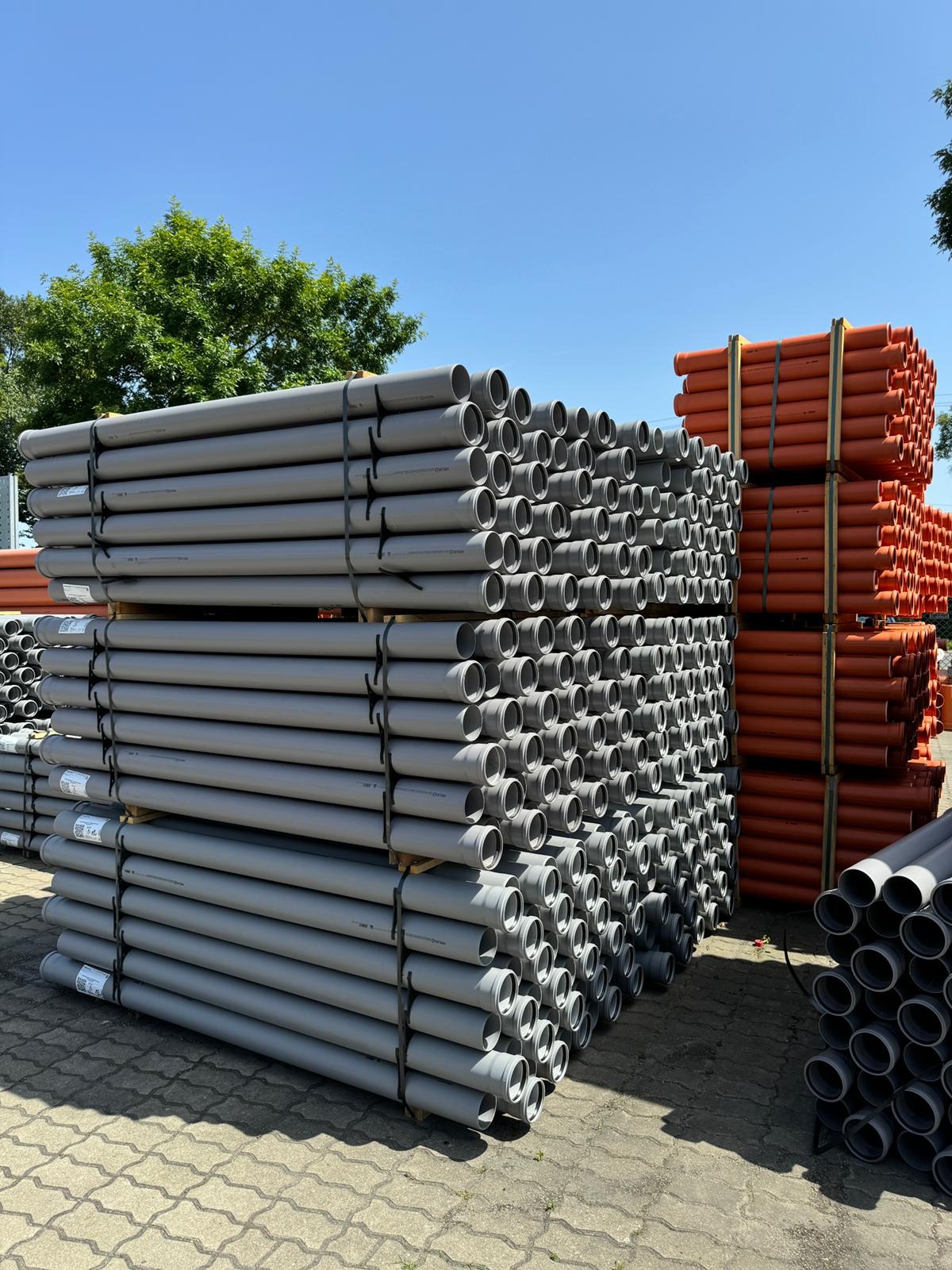 Sztaple szarych i pomarańczowych rur PCV na paletach, widoczne na zewnątrz w słoneczny dzień. Rury ułożone równolegle, tworzą regularny wzór.