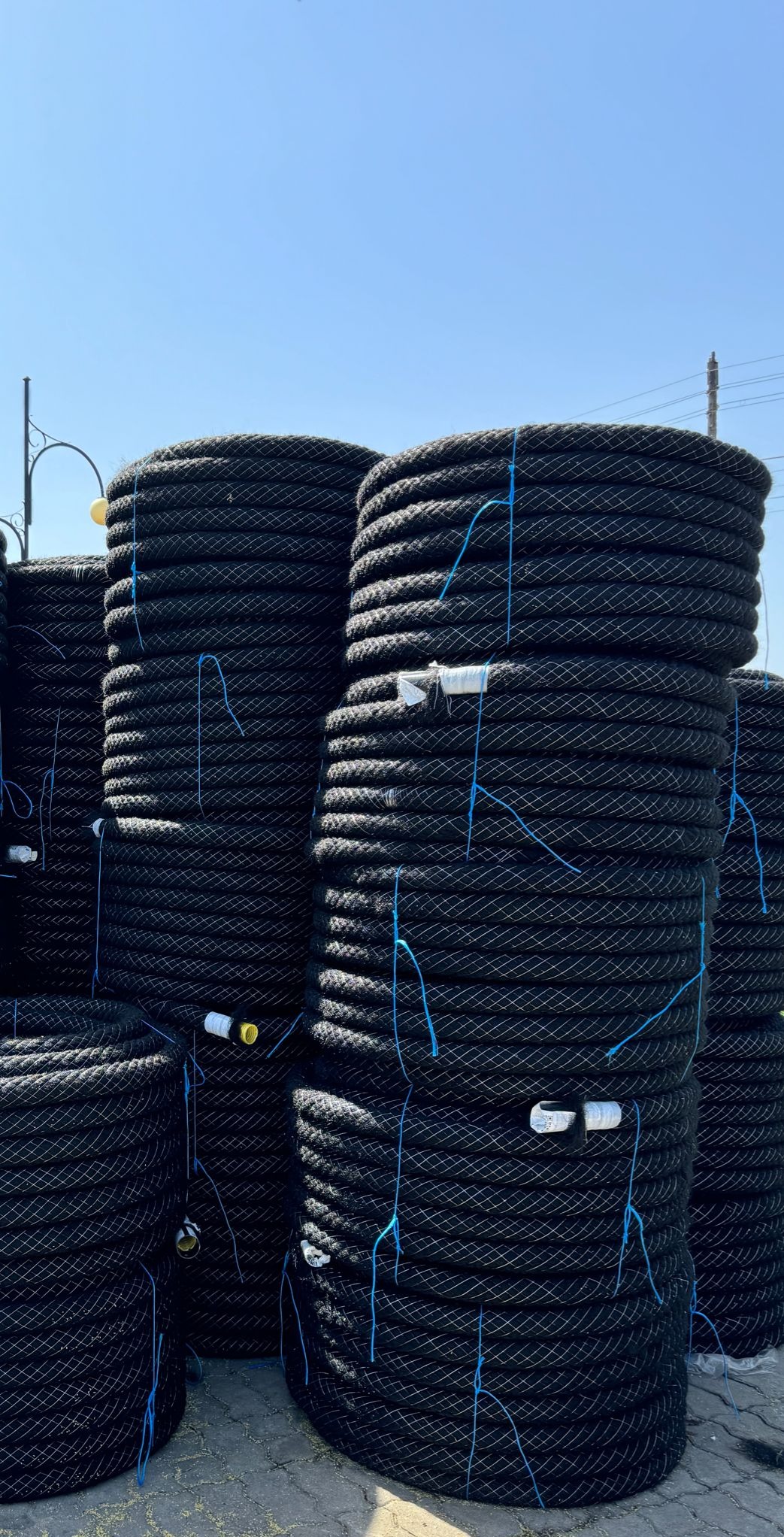 Stosy czarnych rur drenarskich, spięte niebieskim sznurkiem, na zewnątrz w słoneczny dzień. Widoczne studzienki i elementy systemu odwodnień.