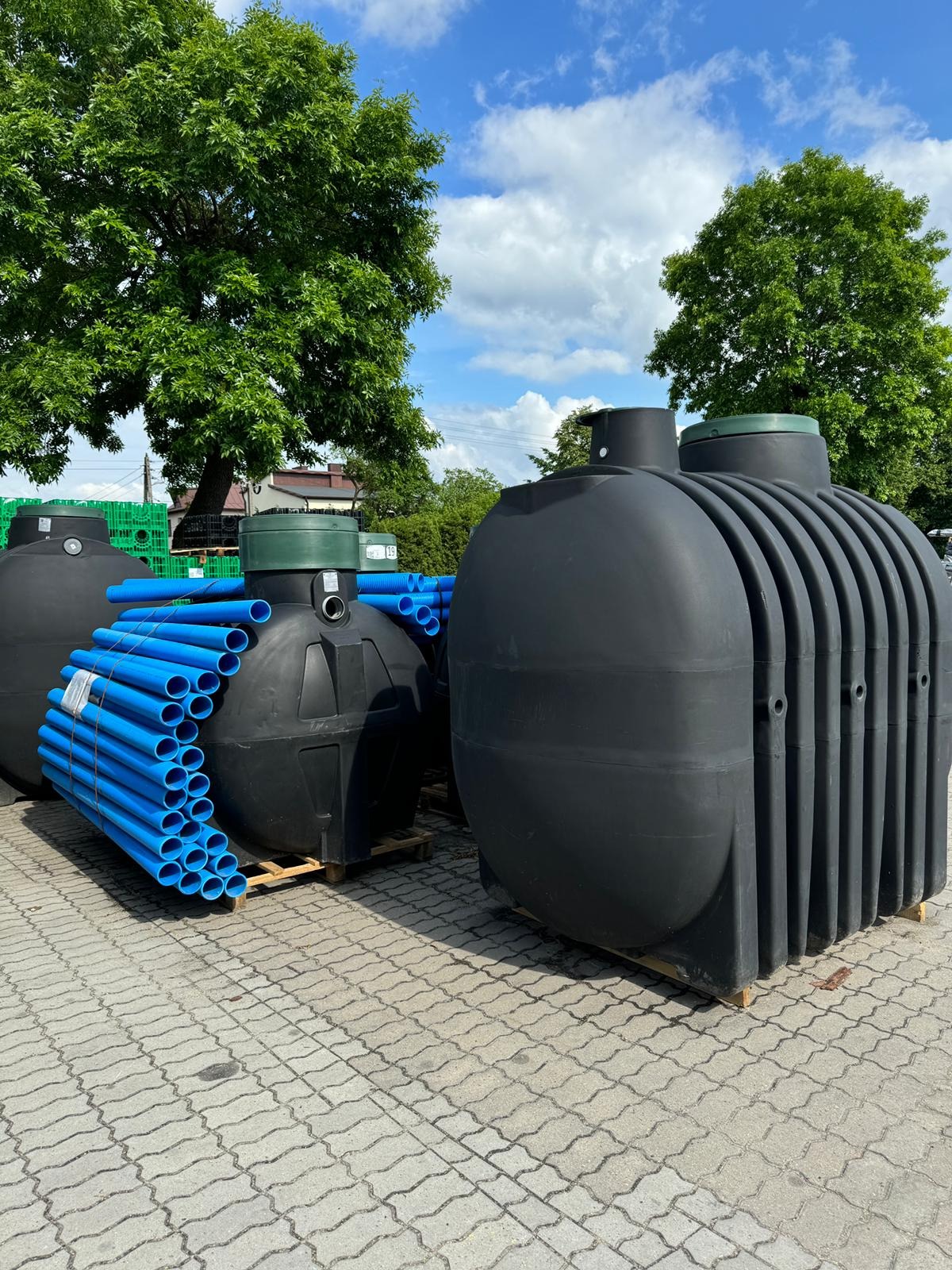 Czarne zbiorniki na paletach i niebieskie rury na zewnątrz, w tle zieleń drzew i błękitne niebo z chmurami. Elementy systemów odwodnień i oczyszczalni.