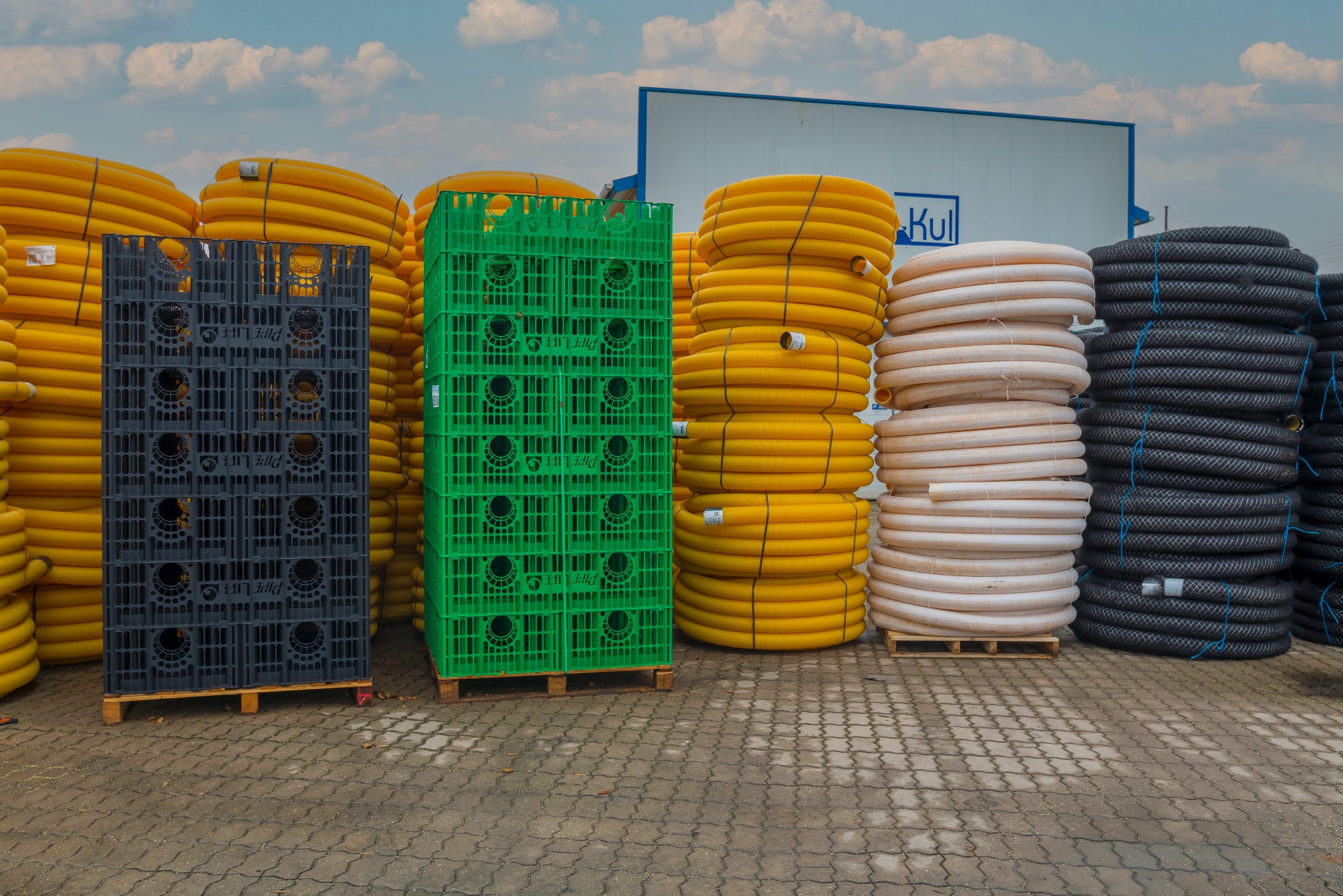 Magazyn rur drenarskich w rolkach, ułożonych obok siebie w stosach, w kolorach: żółtym, beżowym i czarnym. Dodatkowo widoczne czarne i zielone skrzynki.