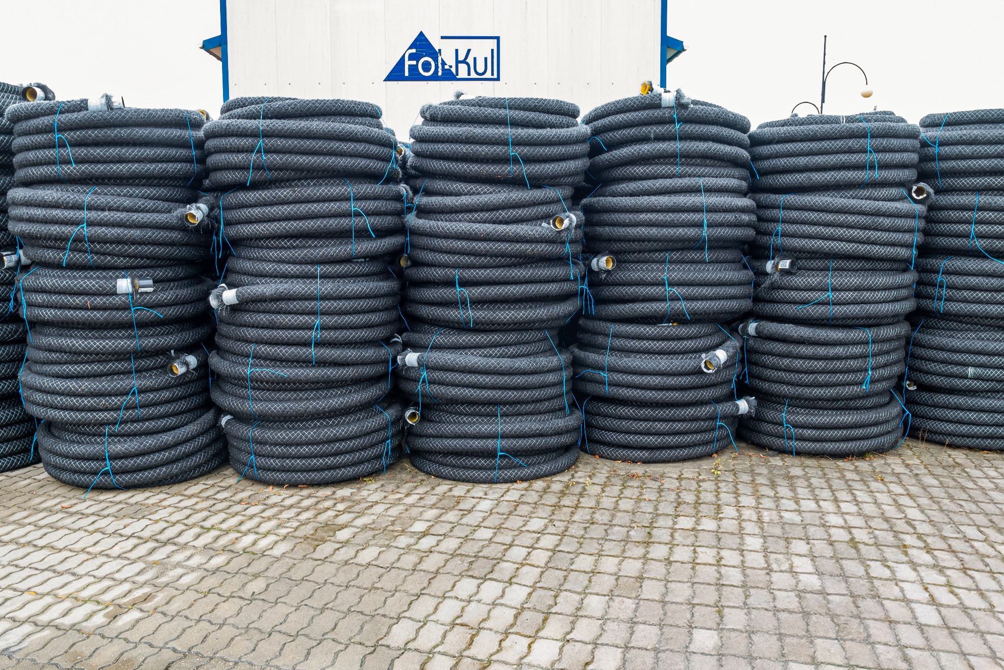 Stosy czarnych rur drenarskich z niebieskim sznurkiem, ułożone na kostce brukowej. W tle biały kontener z logo firmy Fol-Kul. Widok z bliska, oświetlenie dzienne.