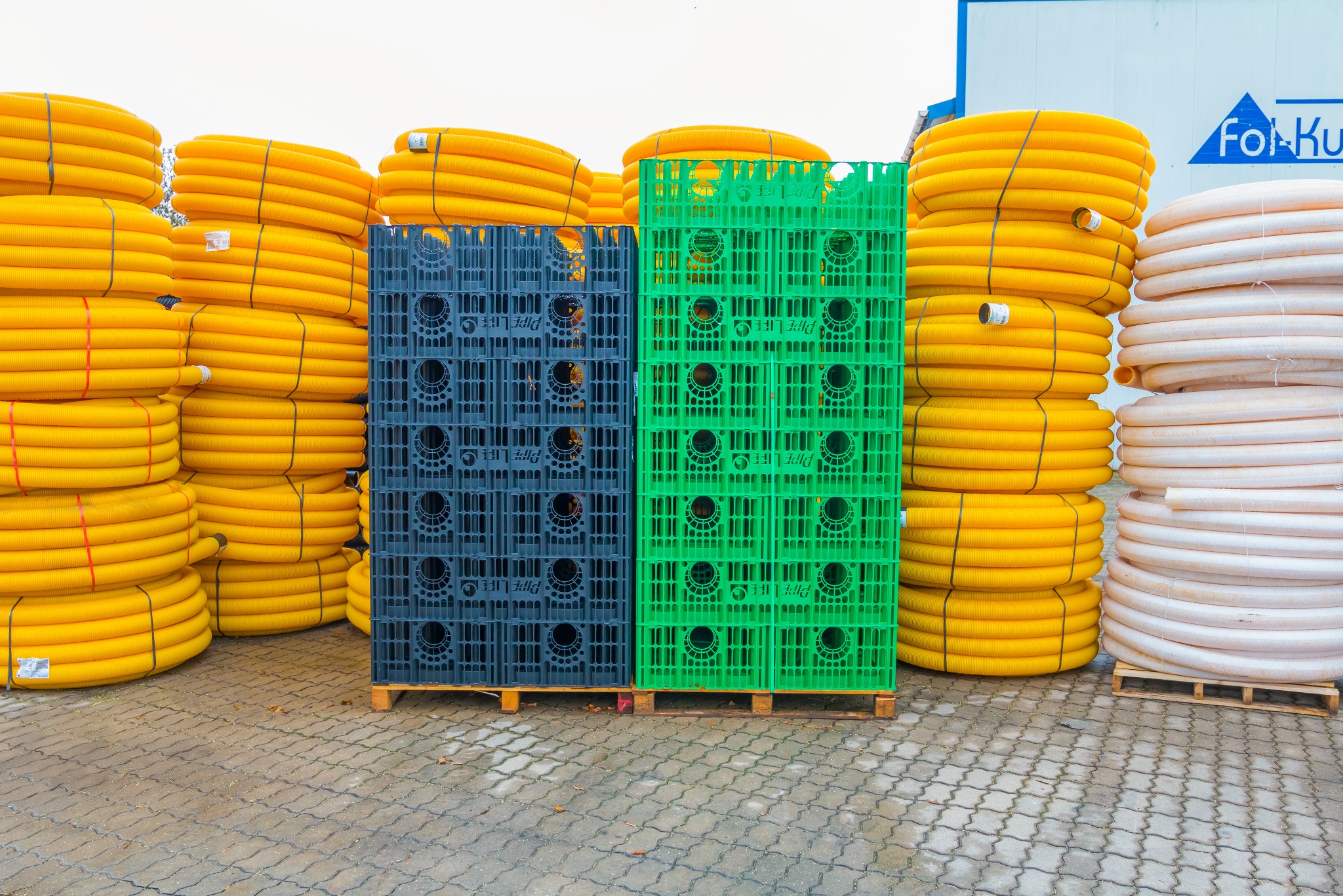 Magazyn rur drenarskich: żółte i białe rury ułożone w stosy obok zielonych i szarych skrzynek na paletach, widok z zewnątrz, jasne oświetlenie.