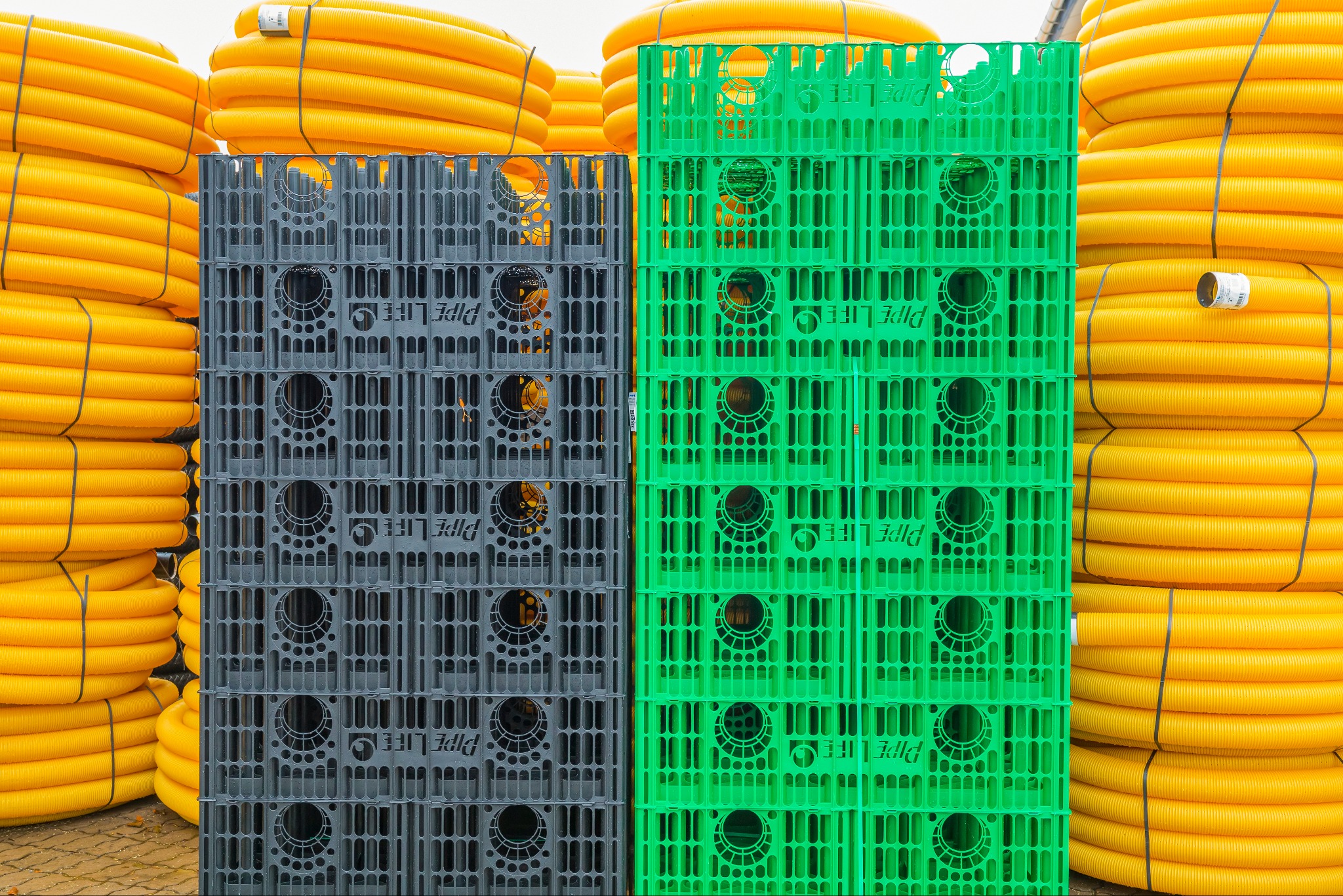 Żółte rury drenażowe zwinięte w kręgi obok stosu szarych i zielonych skrzynek z logo Pipe Life. Elementy składowe systemów odwodnieniowych i oczyszczalni.