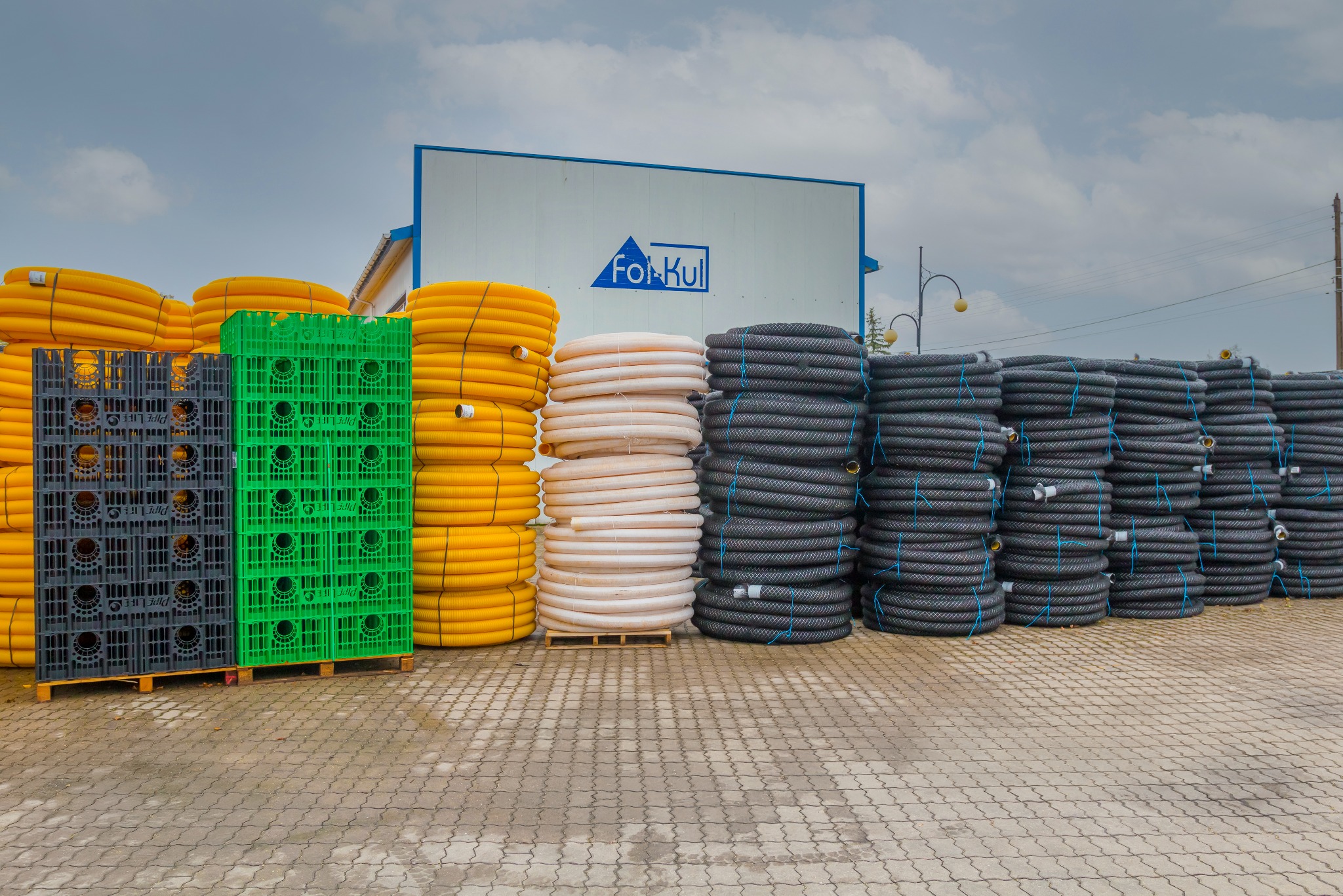 Magazyn rur drenarskich w rolkach, ułożonych kolorami: żółte, beżowe i czarne, spięte niebieskim sznurkiem. Obok stoją skrzynki z tworzywa sztucznego: szare i zielone.