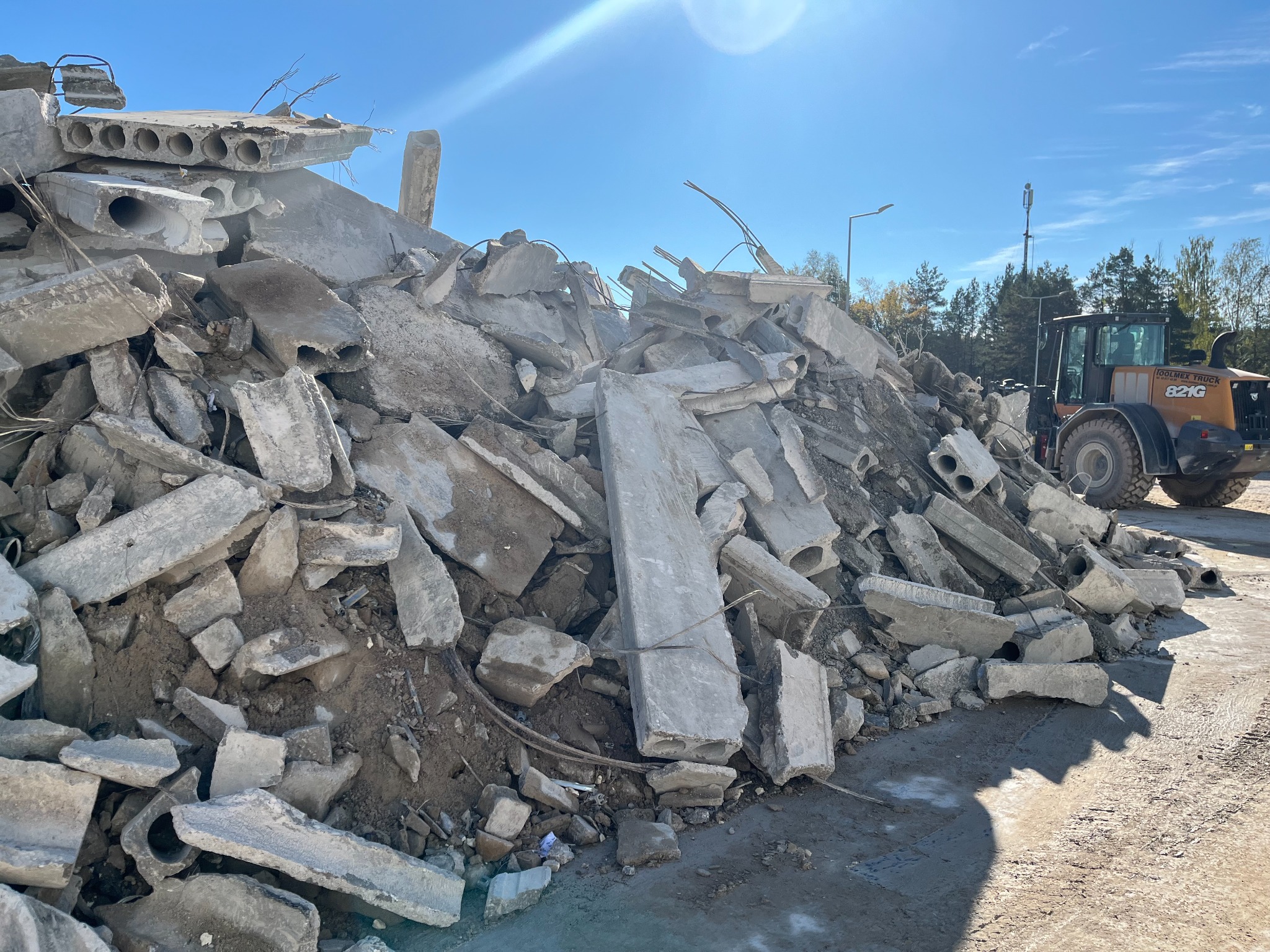 Stos gruzu betonowego z widocznymi fragmentami płyt i zbrojeń, na tle błękitnego nieba i zieleni drzew, z koparką w tle. Widok po rozbiórce.