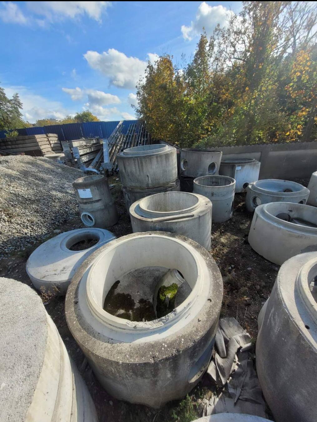 Betonowe kręgi studni na placu składowym, widoczne ślady użytkowania, mech wewnątrz jednego z kręgów, w tle drzewa i błękitne niebo z chmurami.