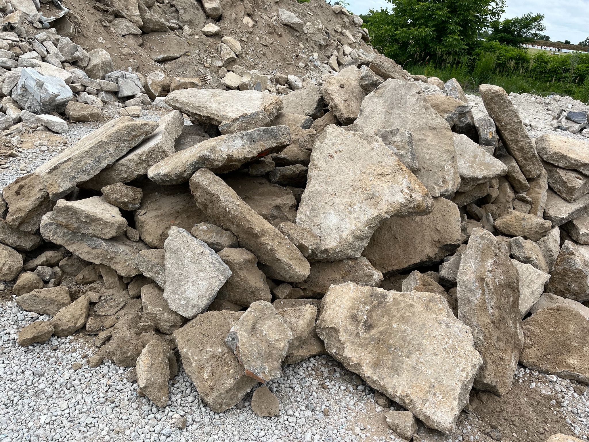 Stos gruzu betonowego z widocznymi fragmentami zbrojenia, ułożony na żwirowym podłożu, z zielenią w tle. Widok z bliska, oświetlenie dzienne.