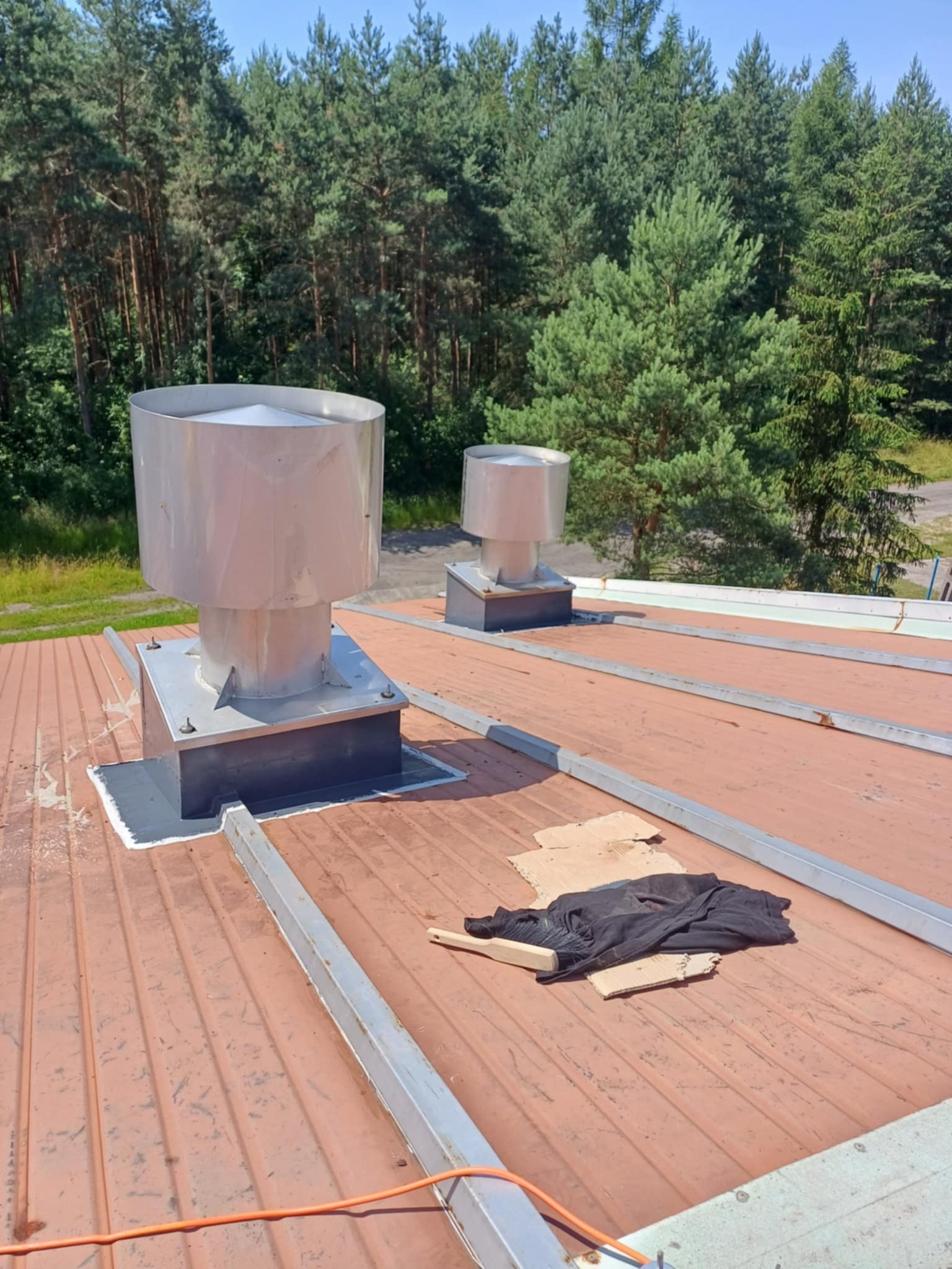 Dwa metalowe kominy wentylacyjne na pomarańczowym dachu z widocznymi uszczelnieniami i lasem w tle. Na dachu leżą kartony i czarna szmata.