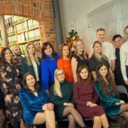 Zdjęcie grupowe zespołu biura na tle ceglanej ściany z regałem pełnym segregatorów i choinki. Uśmiechnięci pracownicy w różnorodnych strojach pozują do zdjęcia.