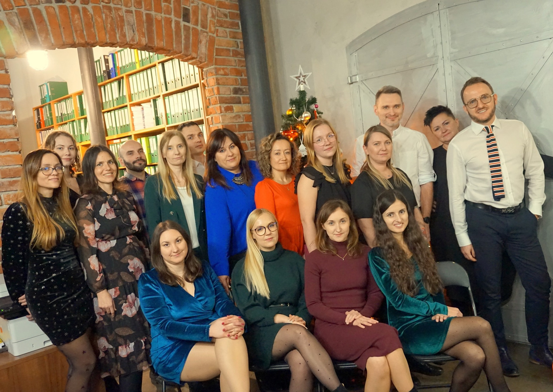 Zdjęcie grupowe zespołu biura na tle ceglanej ściany z regałem pełnym segregatorów i choinki. Uśmiechnięci pracownicy w różnorodnych strojach pozują do zdjęcia.