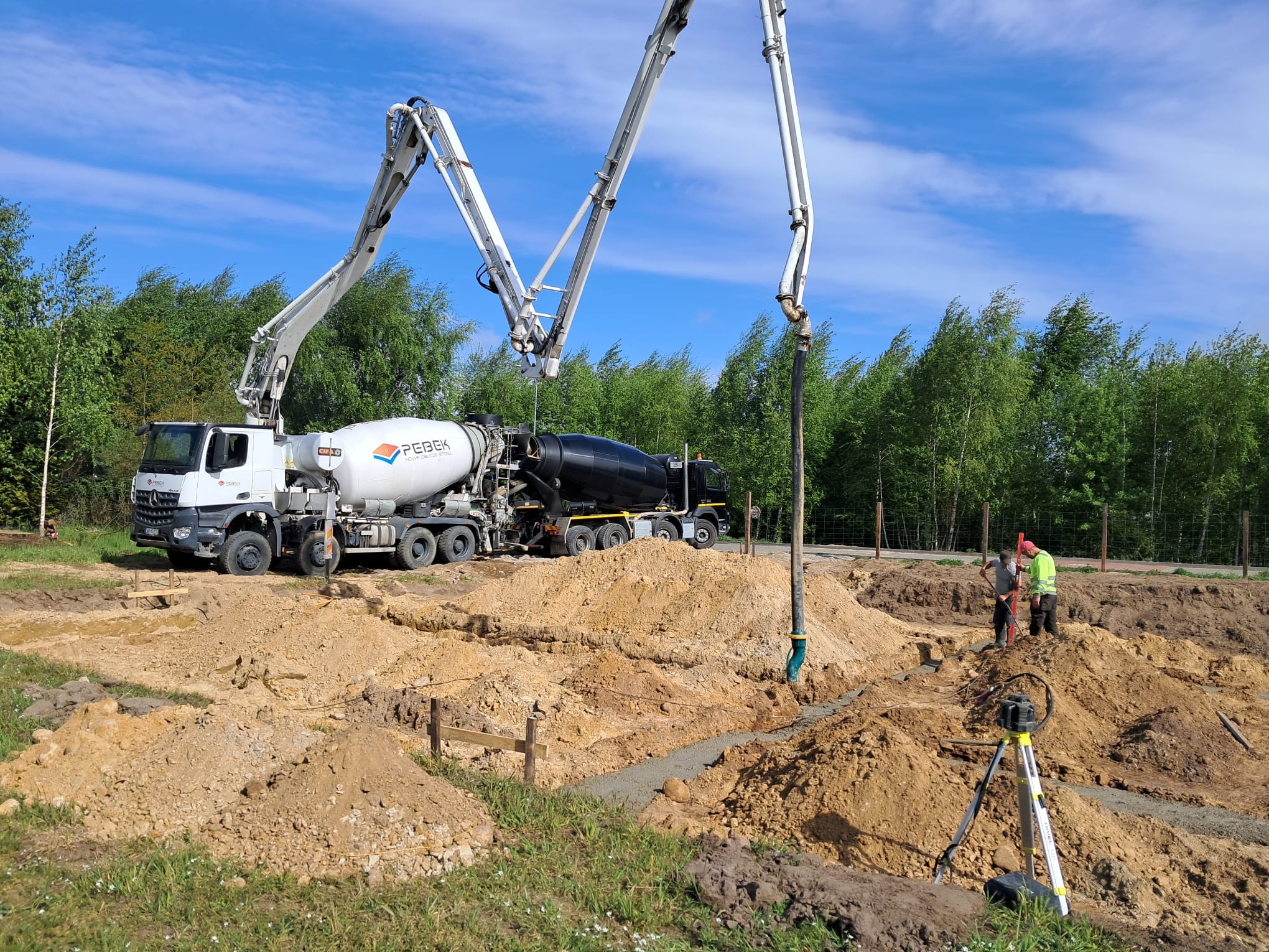 Wylewanie fundamentów pod dom jednorodzinny z użyciem pompy do betonu. Widoczni pracownicy i niwelator laserowy. Świeży beton w wykopie.