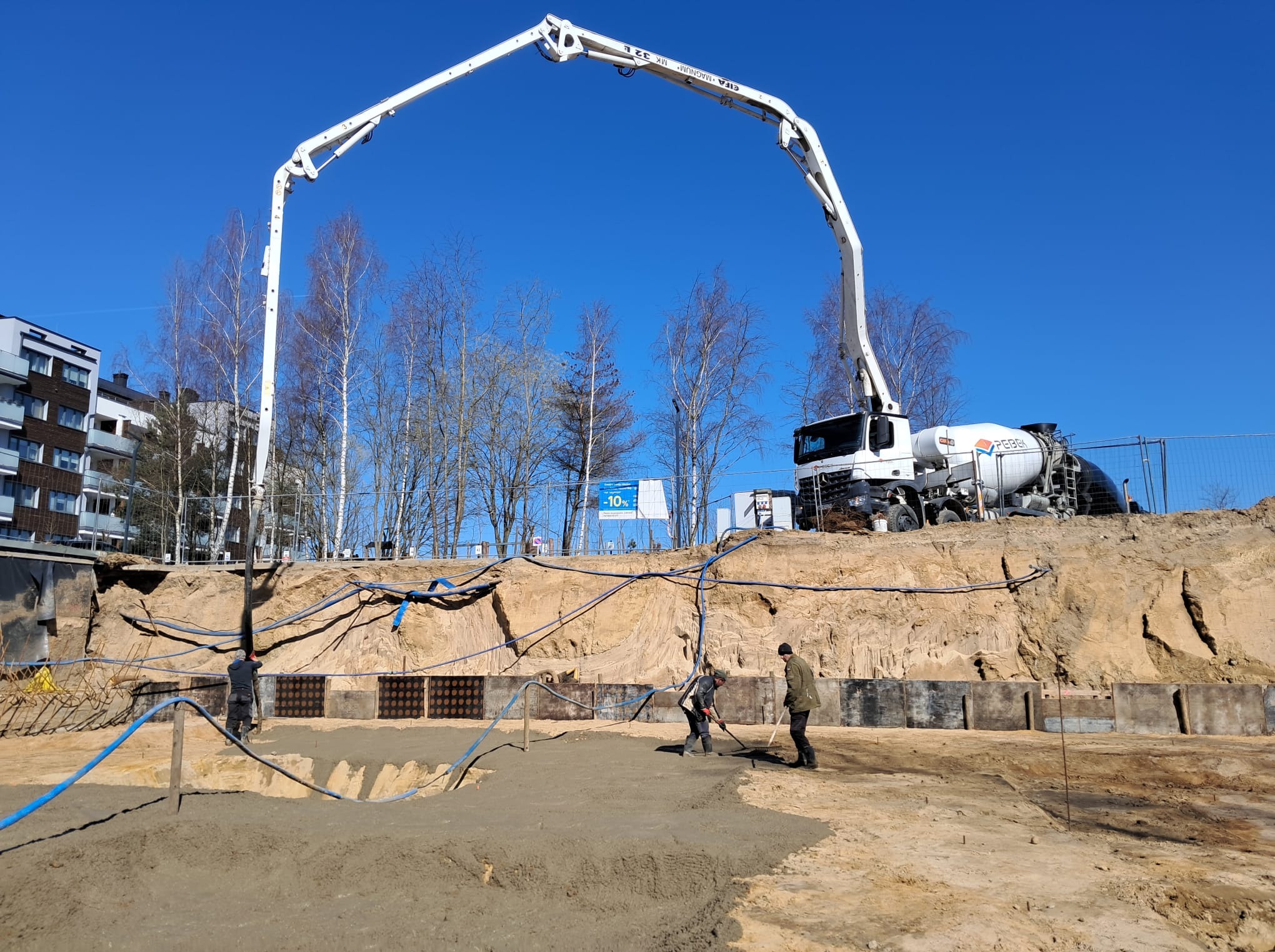 Wylewanie betonu na budowie z użyciem pompy do betonu. Widoczni pracownicy wyrównujący powierzchnię. W tle budynek mieszkalny i błękitne niebo.