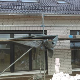 Waldi Bau - Wznoszenie jasnej, murowanej ściany domu z widocznymi oknami i rusztowaniem. Część okna zasłonięta plandeką. Na rusztowaniu butelka wody. Widoczny fragment dachu.