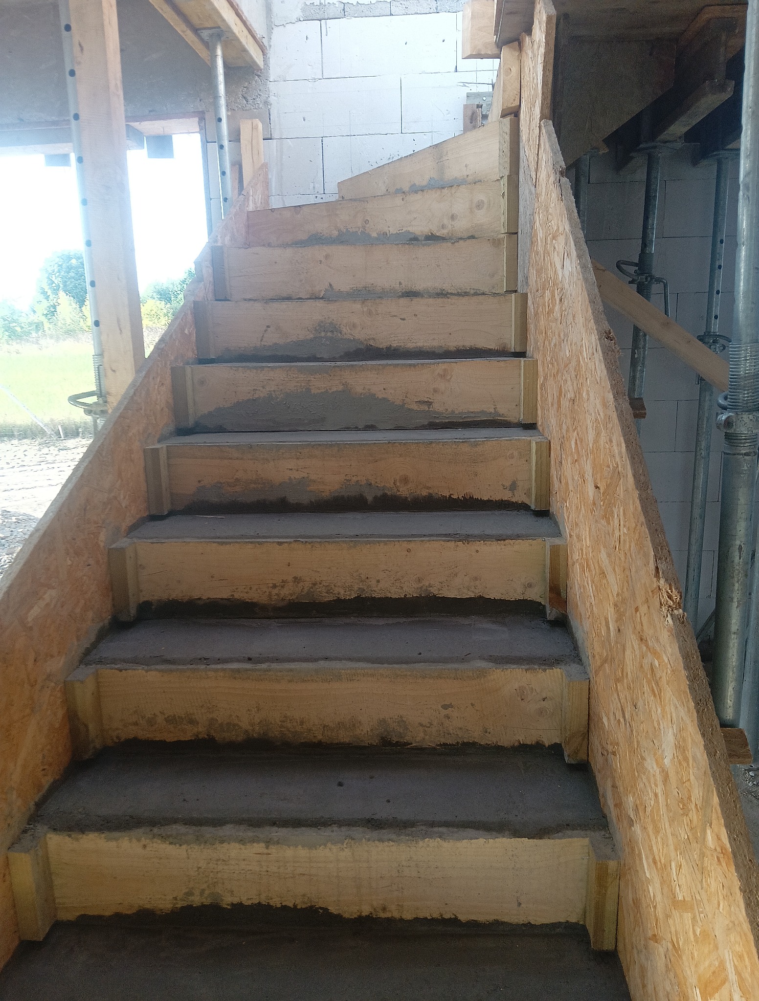 Surowe schody betonowe w drewnianym szalunku na budowie, widoczne podpory i bloczki. Etap wylewania stopni, surowy stan, widok z dołu.