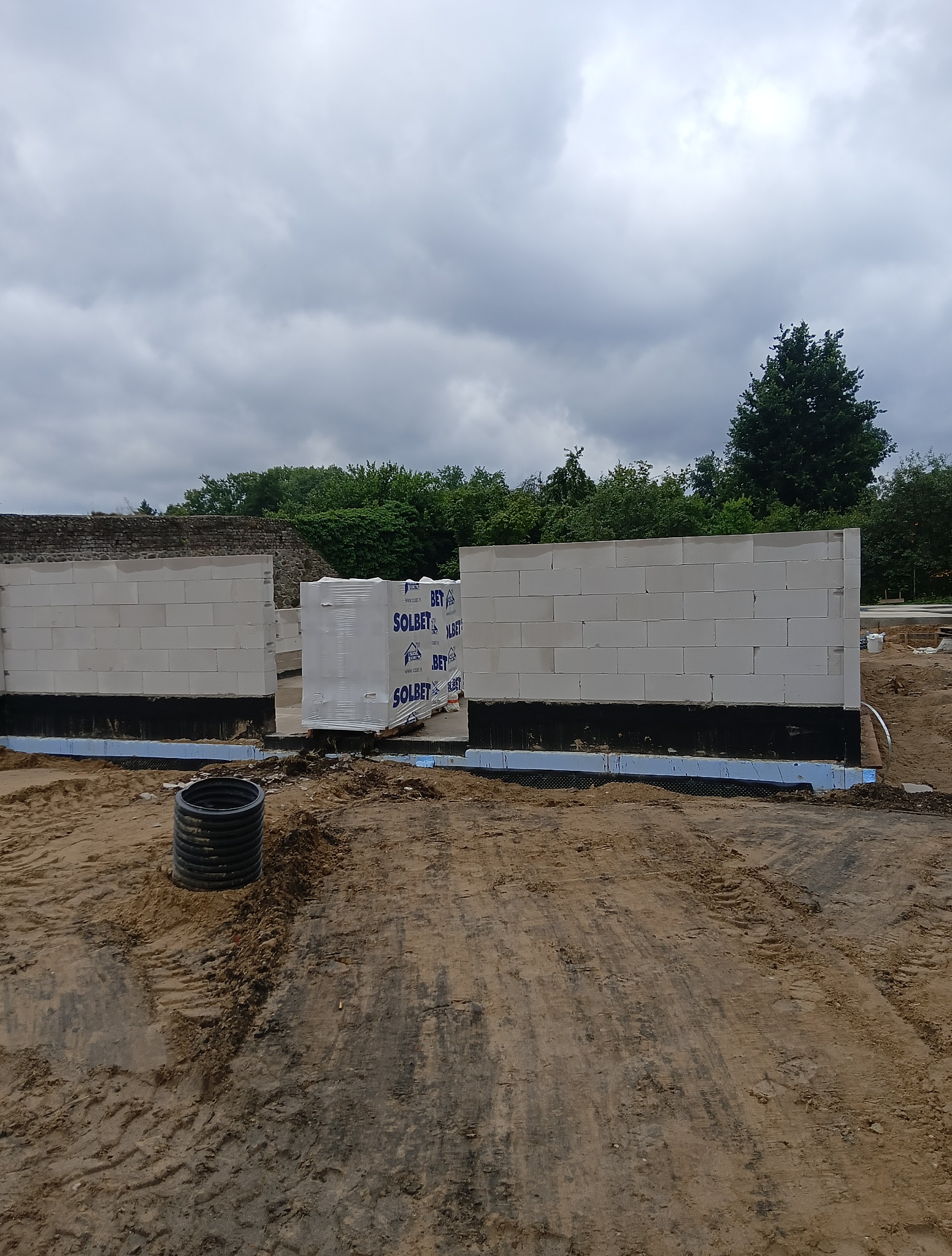 Wznoszone ściany z betonu komórkowego Solbet na fundamencie z izolacją przeciwwilgociową. Częściowo wymurowane ściany, paleta bloczków na placu budowy. Pochmurne niebo.