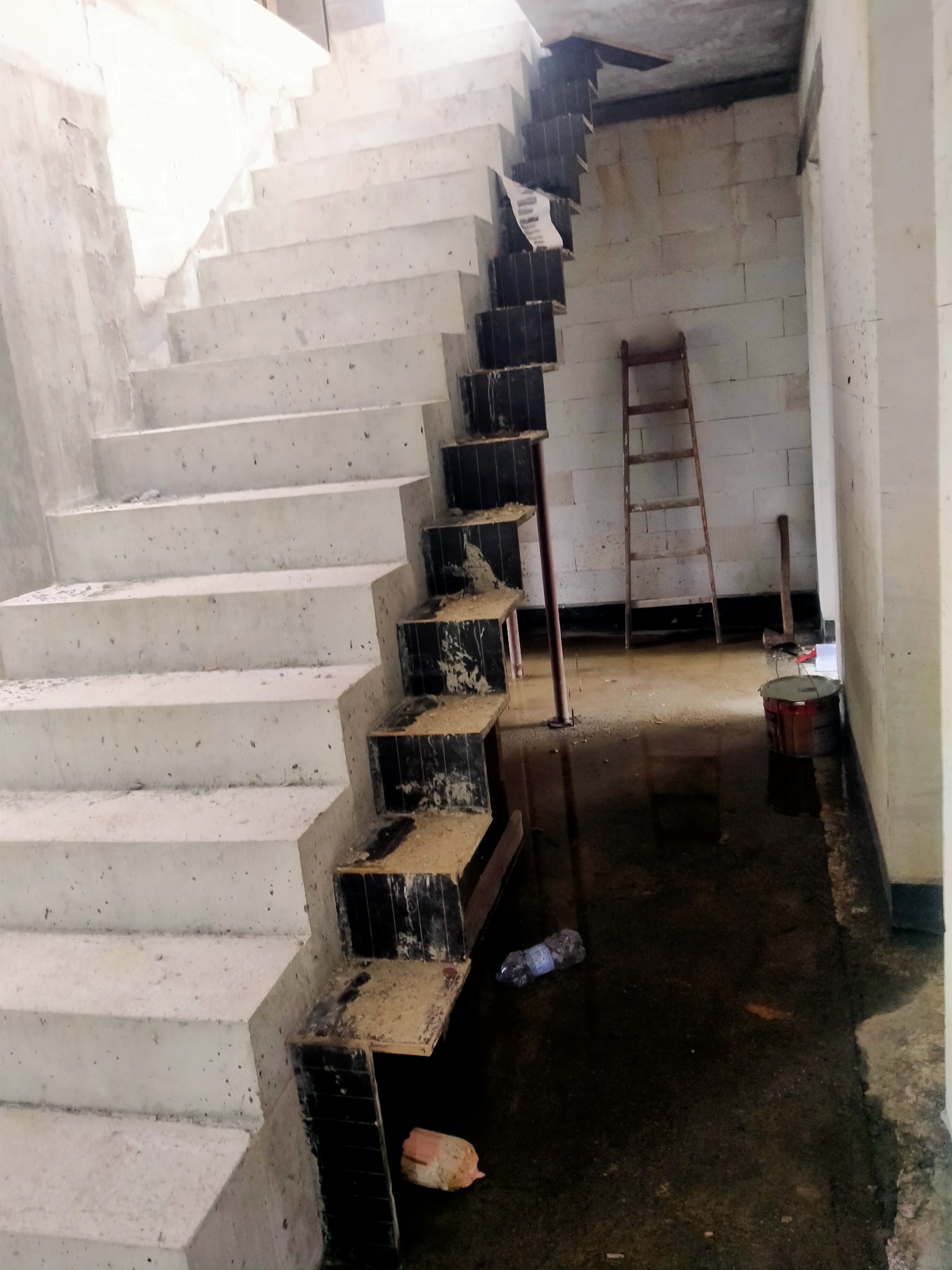 Surowe betonowe schody w trakcie budowy, z widocznymi elementami szalunku i mokrą podłogą. W tle ściana z bloczków i drabina.