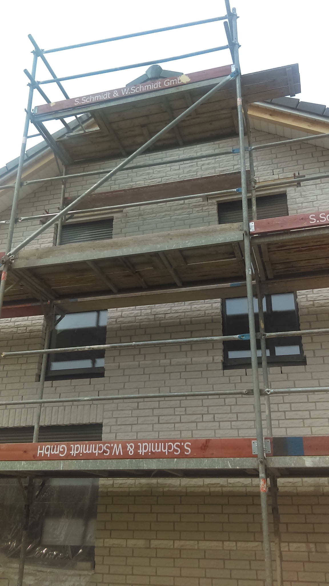 Ściana domu z jasnej cegły w trakcie budowy, widoczna z rusztowania z nazwą firmy S.Schmidt & W.Schmidt GmbH. Częściowo widoczne okna z roletami.