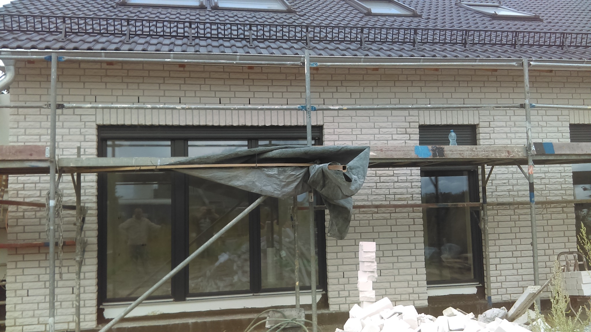 Wznoszenie jasnej, murowanej ściany domu z widocznymi oknami i rusztowaniem. Część okna zasłonięta plandeką. Na rusztowaniu butelka wody. Widoczny fragment dachu.