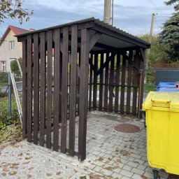 TAB Mateusz Ciask - Drewniana wiata śmietnikowa z metalowym dachem na posesji, obok żółty kontener na śmieci. Widoczne kostki brukowe i opadłe liście. W tle dom i drzewa.