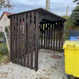 Drewniana wiata śmietnikowa z metalowym dachem na posesji, obok żółty kontener na śmieci. Widoczne kostki brukowe i opadłe liście. W tle dom i drzewa.