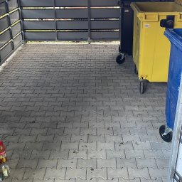 TAB Mateusz Ciask - Zadaszone miejsce na pojemniki na odpady z szarej kostki brukowej. Widoczne pojemniki w kolorach: żółtym, niebieskim i czarnym. Ślady po płynach na kostce.