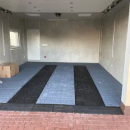 Wykończony garaż z szarą podłogą z kostki brukowej i dwoma czarnymi pasami na koła. Widoczna brama garażowa, ściany z oknem i karton. Czysto i schludnie.