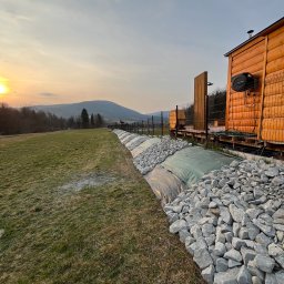 TAB Mateusz Ciask - Wykończony brzeg trawnika z kamieniami i geowłókniną, widok na drewniany budynek i góry w tle, oświetlone zachodzącym słońcem.