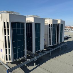BRIMAN SPÓŁKA Z OGRANICZONĄ ODPOWIEDZIALNOŚCIĄ - Rząd pięciu białych jednostek zewnętrznych klimatyzacji Fujitsu AIRSTAGE INVERTER na płaskim dachu, zamontowanych na metalowej konstrukcji, pod błękitnym niebem z widokiem na miasto.