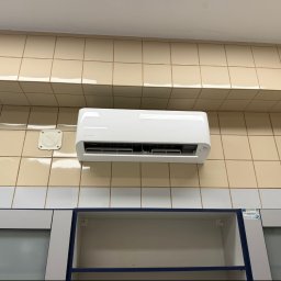 BRIMAN SPÓŁKA Z OGRANICZONĄ ODPOWIEDZIALNOŚCIĄ - Biała jednostka klimatyzacji Rotenso z cyfrowym wyświetlaczem 21.0, zamontowana na ścianie z beżowych i jasnych płytek, nad otwartą szafką biurową.