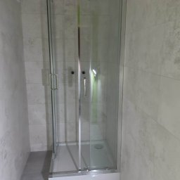 Jarosław Aniołowski - Kabina prysznicowa z przesuwnymi drzwiami, wykończona jasnymi płytkami w łazience. Widoczny odpływ liniowy i minimalistyczne uchwyty.