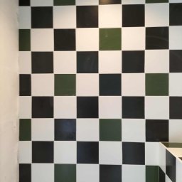 Jarosław Aniołowski - Ściana wyłożona błyszczącymi, kwadratowymi płytkami w szachownicę: białe, czarne i oliwkowe. Fragment narożnika z identycznym wzorem płytek.