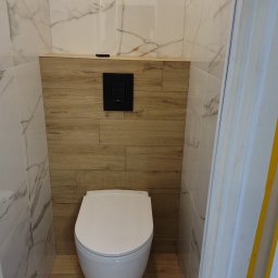 Jarosław Aniołowski - Nowoczesna toaleta w wykończonej łazience: biała ceramika, marmurowe i drewnopodobne płytki, czarny panel sterujący. Widok z perspektywy drzwi.