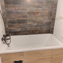 Jarosław Aniołowski - Wykończona wanna z obudową imitującą drewno i baterią w ciemnym kolorze. Na ścianie płytki w odcieniach brązu i szarości. Widoczny otwór rewizyjny.