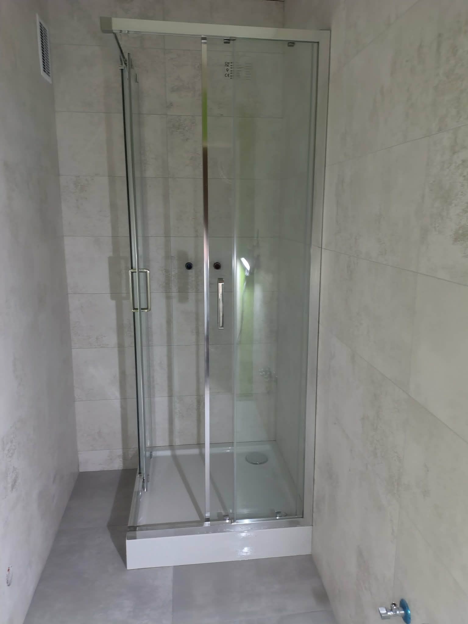 Kabina prysznicowa z przesuwnymi drzwiami, wykończona jasnymi płytkami w łazience. Widoczny odpływ liniowy i minimalistyczne uchwyty.