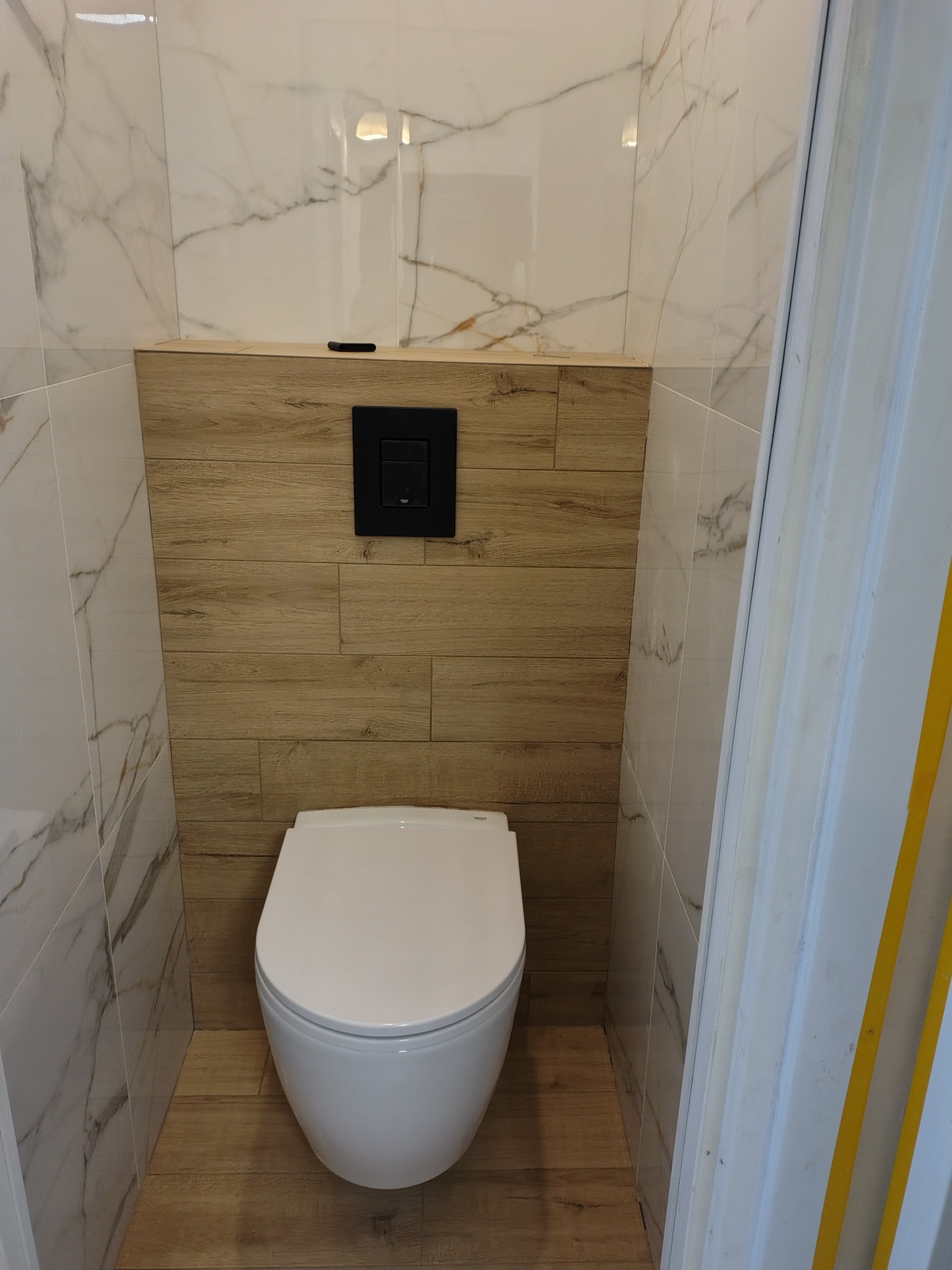 Nowoczesna toaleta w wykończonej łazience: biała ceramika, marmurowe i drewnopodobne płytki, czarny panel sterujący. Widok z perspektywy drzwi.