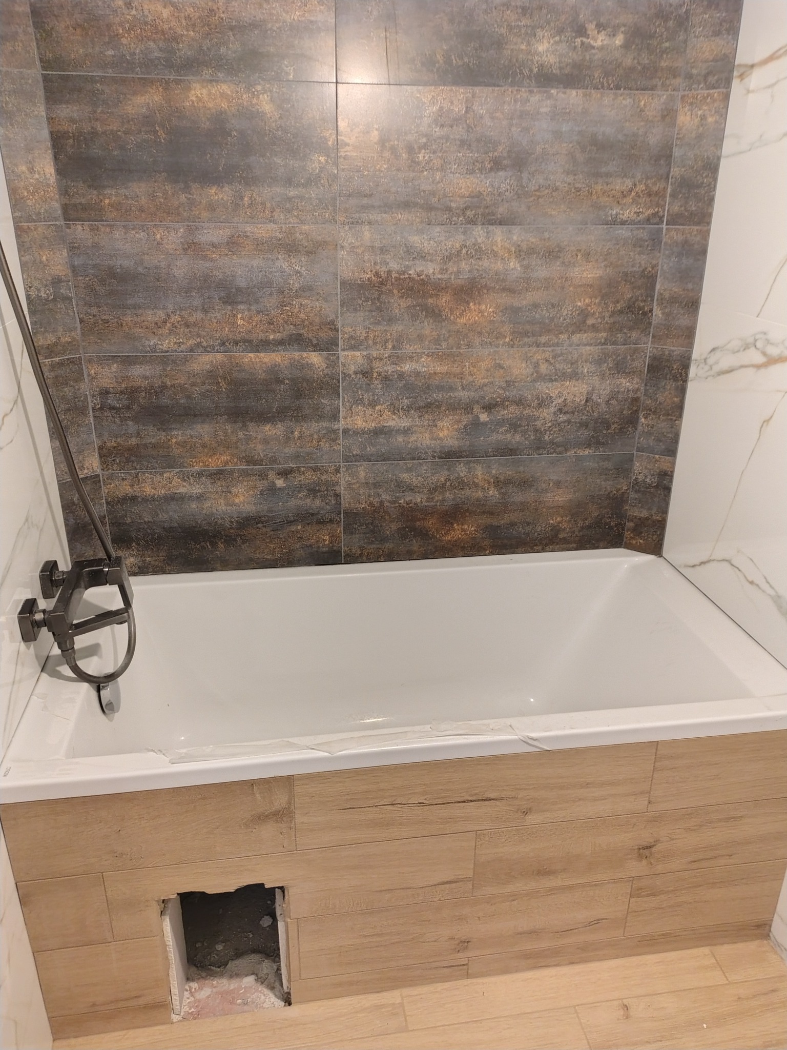 Wykończona wanna z obudową imitującą drewno i baterią w ciemnym kolorze. Na ścianie płytki w odcieniach brązu i szarości. Widoczny otwór rewizyjny.