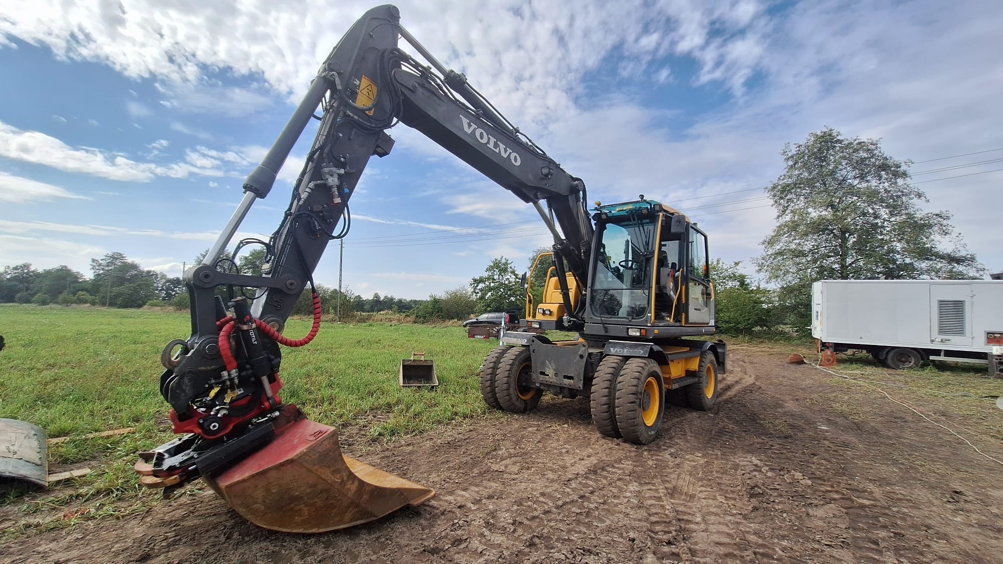 Żółto-czarna koparka Volvo na grząskim terenie, z łyżką skierowaną w dół, gotowa do pracy. W tle zielona łąka i błękitne niebo z chmurami.