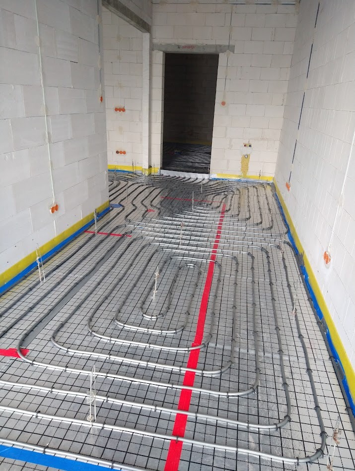 Wnętrze w trakcie montażu ogrzewania podłogowego: rury ułożone na folii z siatką, zabezpieczone ściany, widoczne instalacje elektryczne.