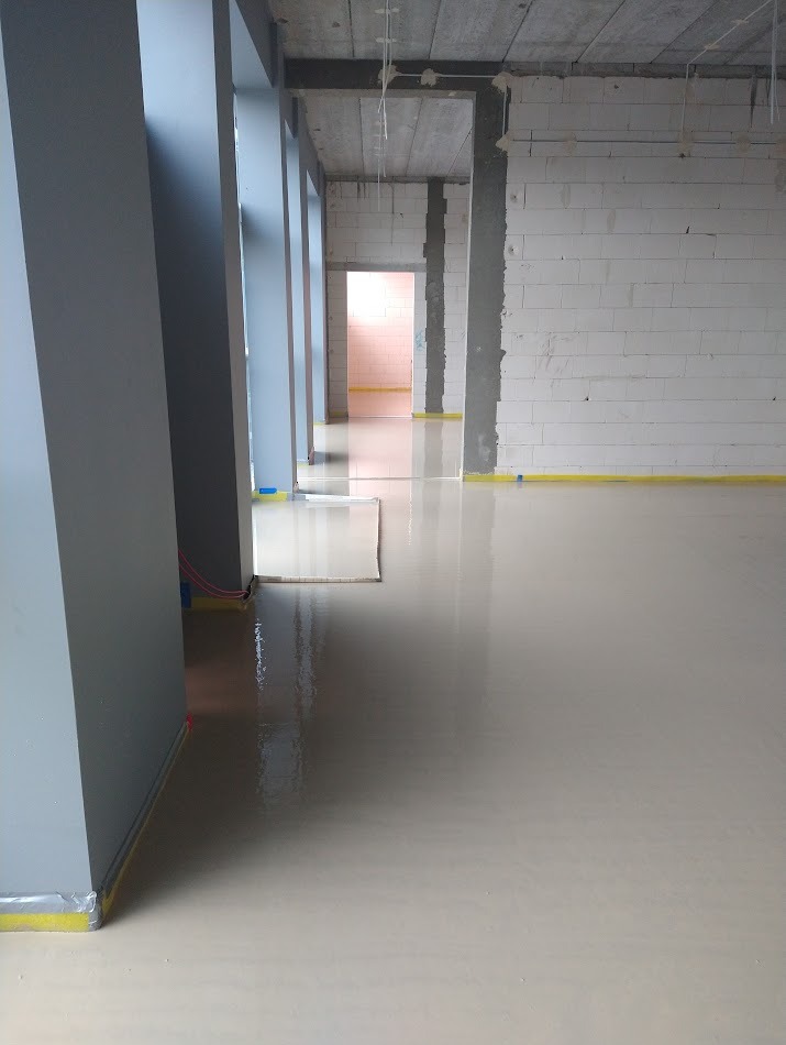 Jasna, gładka posadzka w surowym wnętrzu z białymi ścianami i szarymi filarami. Żółta taśma zabezpieczająca przy ścianach. Widok na korytarz z otwartymi drzwiami.