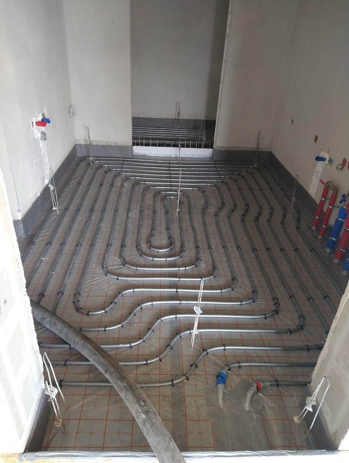 Ułożone rury ogrzewania podłogowego na folii z siatką, przygotowane do zalania. Widoczny wąż do wylewki i fragment ściany z instalacją wodną.