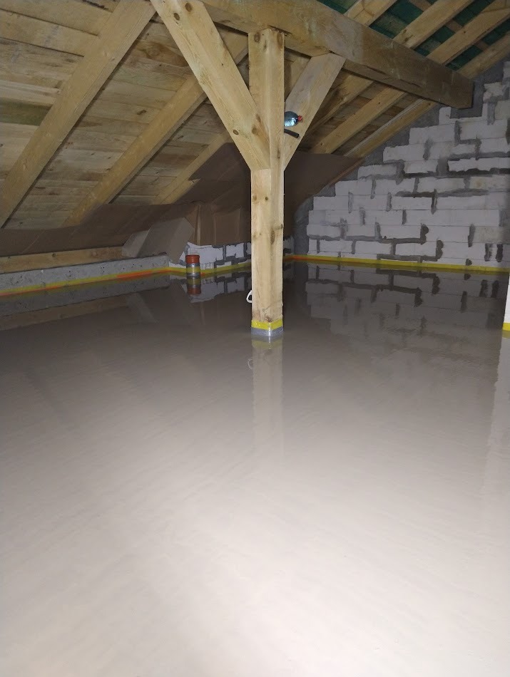 Gładka, jasna posadzka w surowym wnętrzu poddasza z drewnianą konstrukcją dachu i ścianą z bloczków. Żółta taśma oddziela wylewkę od ściany.